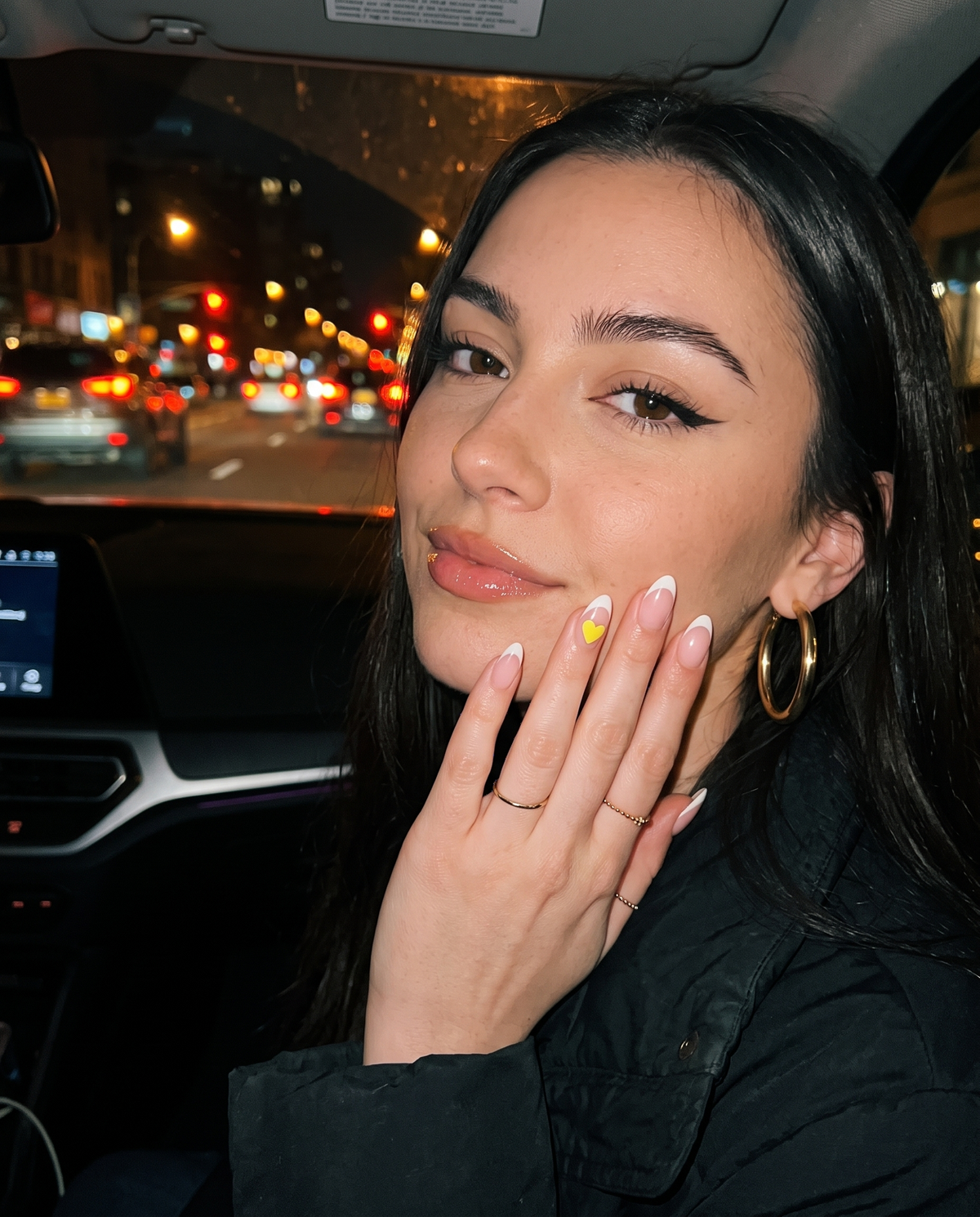 Young woman in a car at night showing a yellow Lumeria heart sticker on her French-tip nail