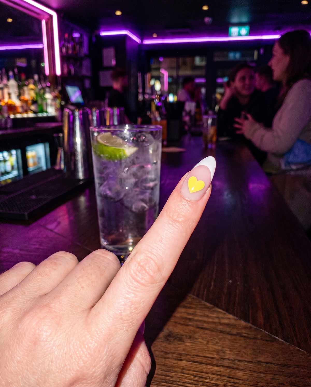 Yellow Lumeria heart sticker on an index nail next to a gin and tonic at a cocktail bar